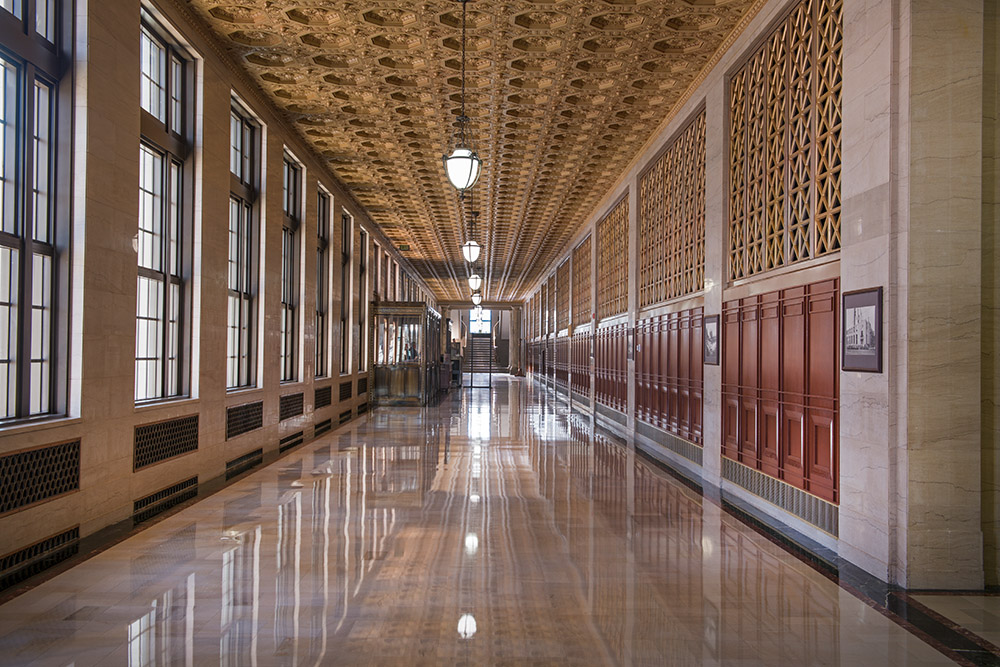 Grand marble-floored hallway with ornate coffered ceiling, tall windows, and wooden paneling in a historic government or institutional building.