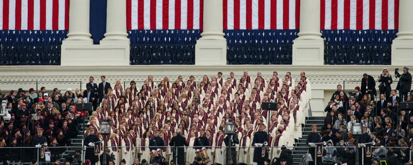 A large choir in white and maroon robes performs on steps before U.S. flags and classical columns at a formal national event.