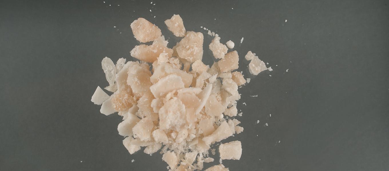 A close-up of irregular, pale beige crystalline fragments scattered on a dark gray surface, likely food-grade or cosmetic-grade crystals.