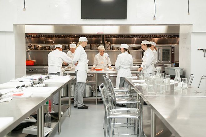 Chefs in white uniforms collaborate in a modern, stainless-steel professional kitchen during a culinary class or workshop.