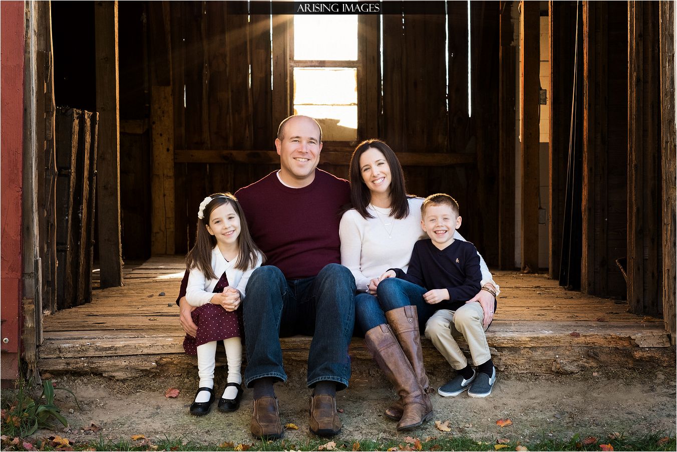 A smiling family of four sits on wooden steps inside a rustic barn, bathed in warm sunlight from a back window.