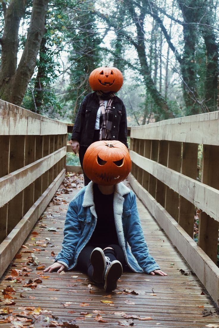 Two people on a wooden bridge wearing carved pumpkin heads, autumn leaves scattered around.