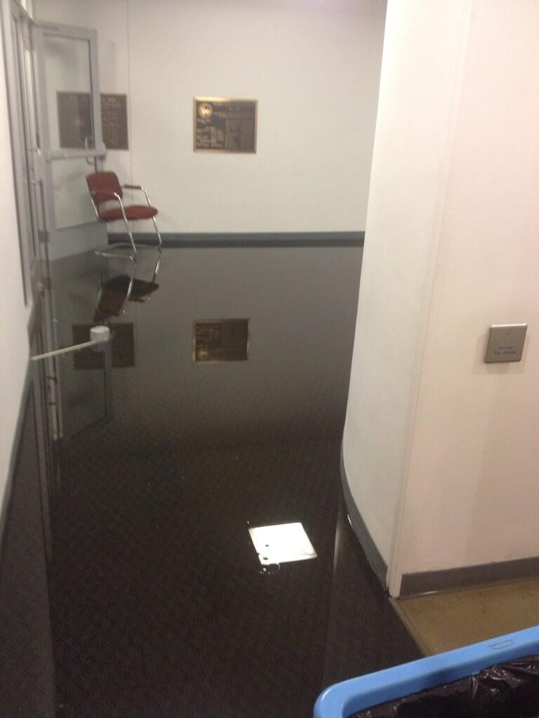 Flooded indoor hallway with reflective water, red chair, wall plaques, and scattered paper on dark floor.