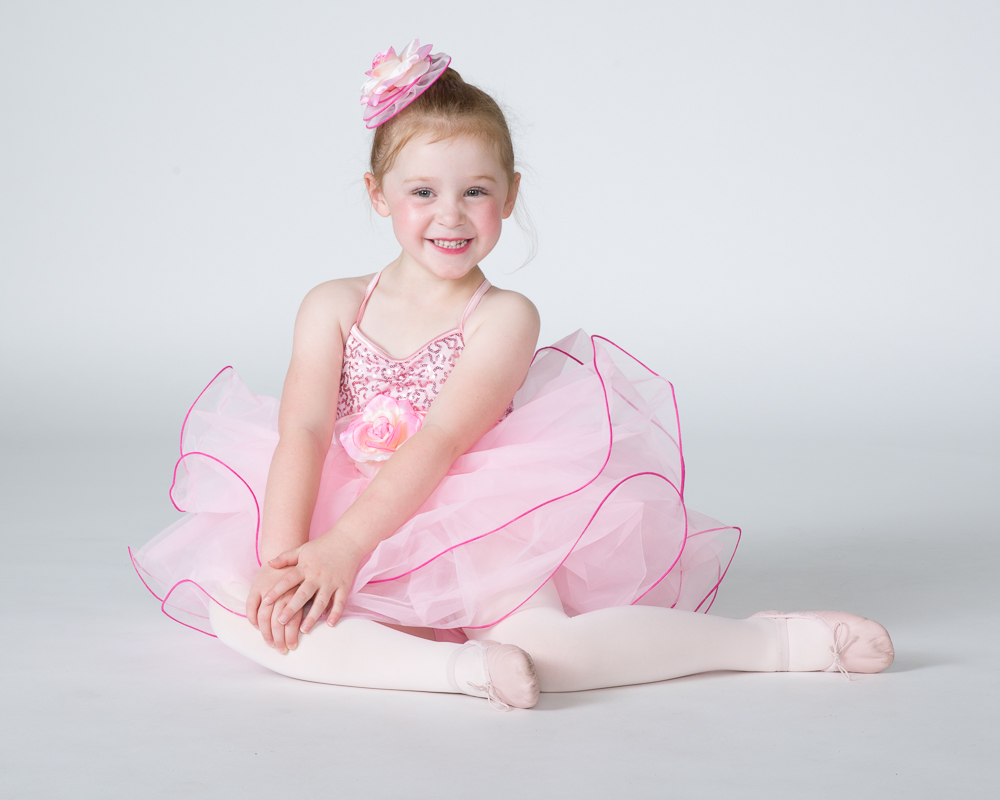 Smiling young ballerina in pink tutu and floral hair accessory, seated on white studio background.