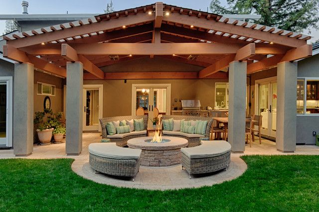 Outdoor patio with wooden pergola, stone fire pit, wicker seating, and lush green lawn at dusk.