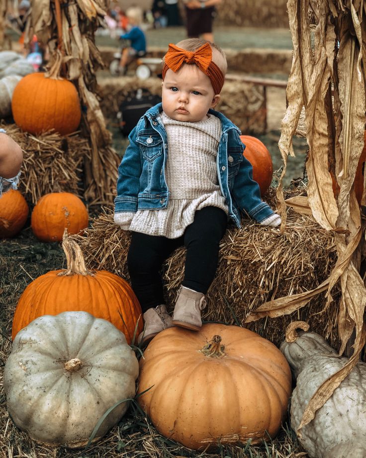Toddler in denim jacket and orange bow sits on hay bale surrounded by pumpkins and corn stalks at autumn farm event.