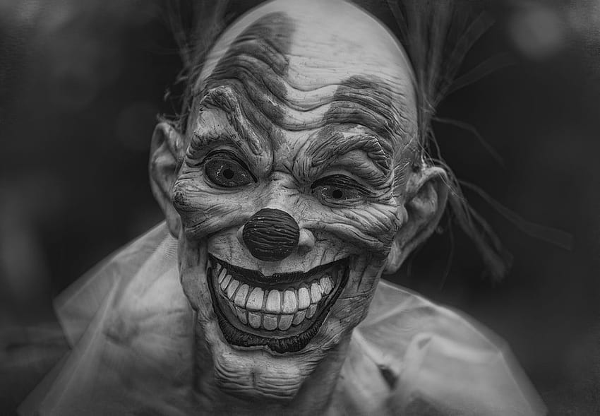 Black-and-white close-up of a grotesque, wrinkled clown mask with exaggerated smile and dark eyes.