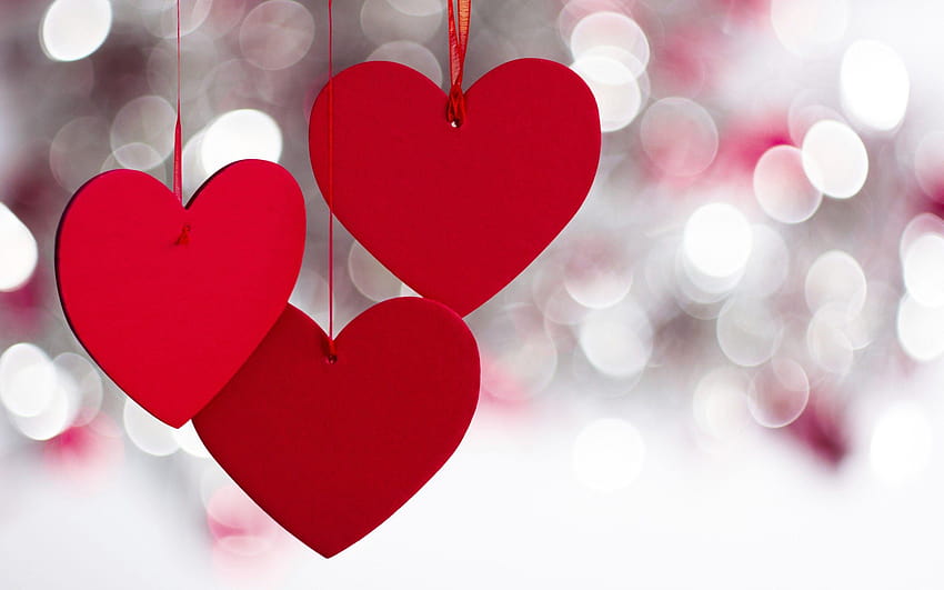 Three red paper hearts hanging from ribbons against a soft bokeh background of white and pink lights.