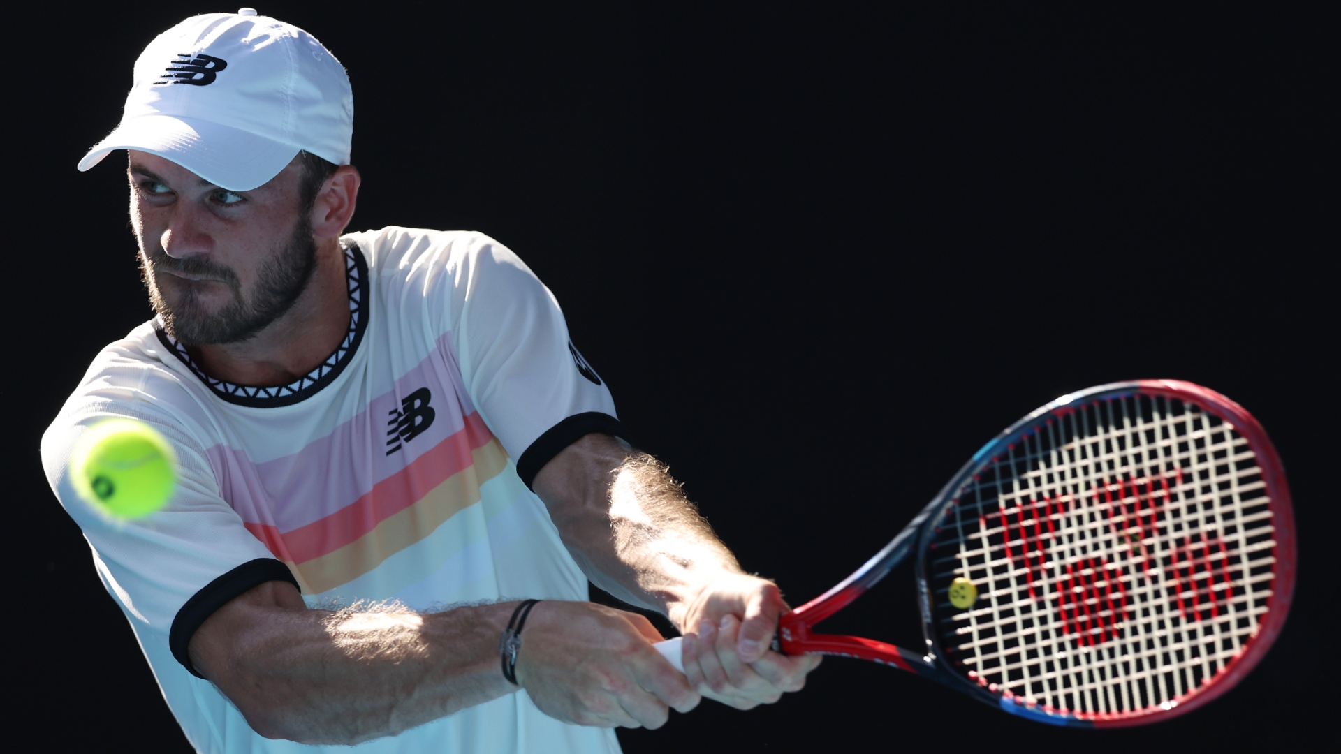 Tennis player in white New Balance outfit mid-swing with Wilson racket, hitting yellow ball against black background.