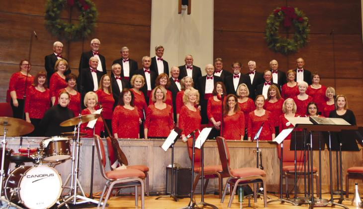 A mixed choir in red and black formal attire poses on stage with instruments, wreaths, and music stands.