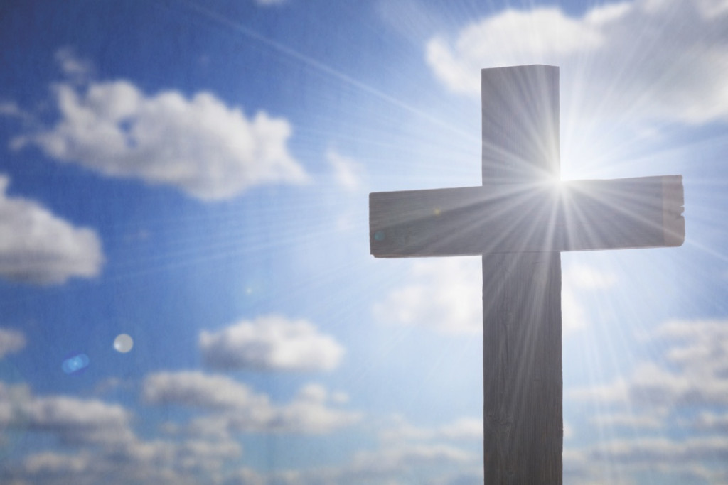 Wooden cross silhouetted against a bright blue sky with sunburst rays and scattered clouds.