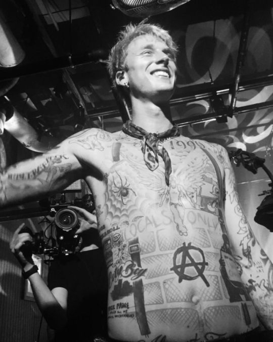 Shirtless man with extensive tattoos smiling under stage lights, photographed in black and white backstage.