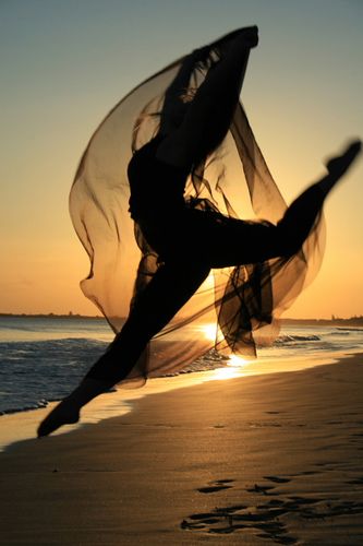 Silhouetted dancer leaping on beach at sunset with flowing fabric and ocean backdrop.