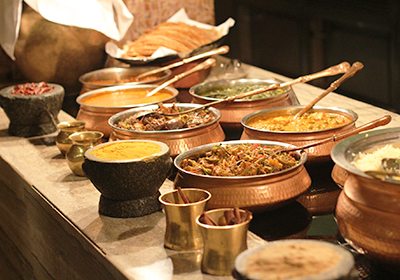 A vibrant Indian buffet spread with copper bowls of curries, chutneys, rice, and breads on a rustic wooden counter.