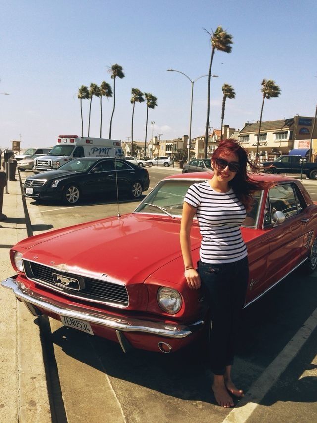 A woman with red hair and sunglasses poses beside a classic red 1960s Ford Mustang on a sunny beachside street lined with palm trees.