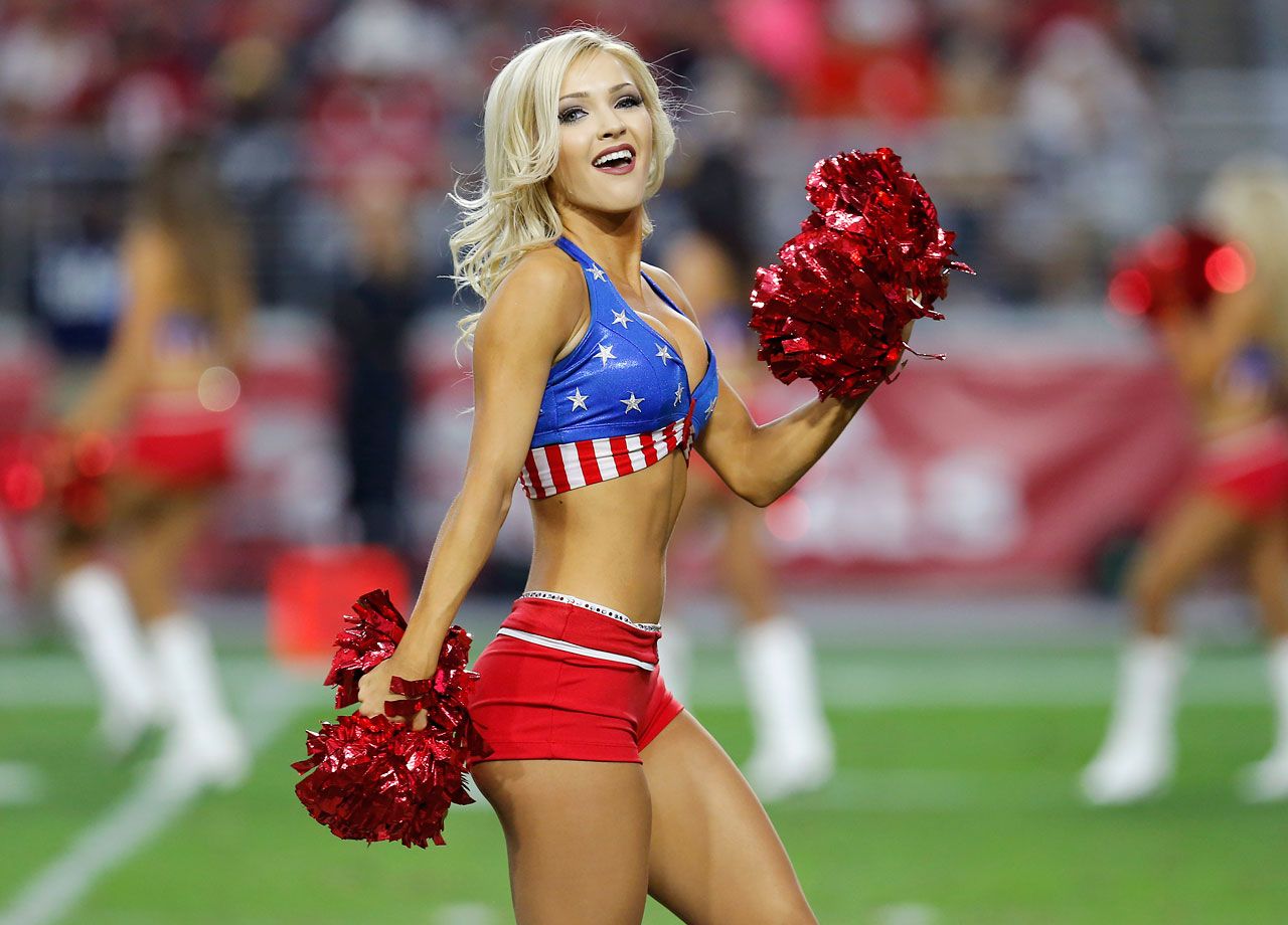 Blonde cheerleader in patriotic blue-and-red uniform with star-spangled top, red pom-poms, performing on football field.