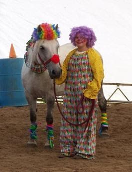 A clown in a colorful outfit and purple wig stands beside a white horse adorned with rainbow hair, noseband, and leg wraps at a circus event.