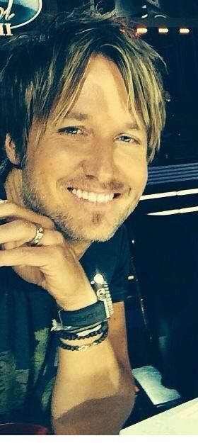 Country singer Keith Urban smiling warmly, wearing bracelets and rings, with "American Idol" logo visible in background.