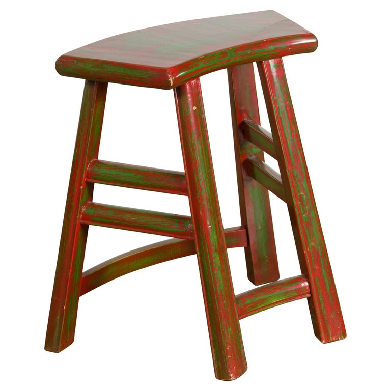 Distressed red-and-green wooden stool with curved seat and A-frame legs, showcasing rustic charm and vintage finish.