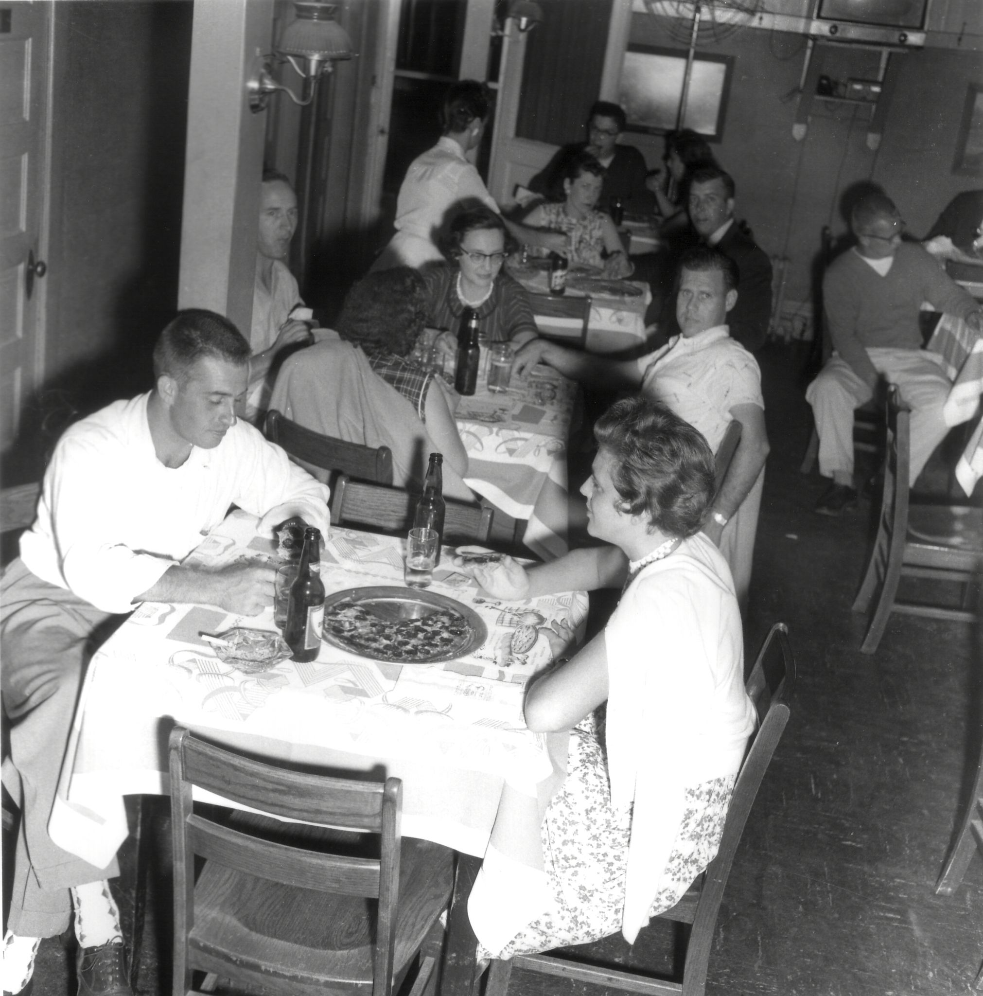 A black-and-white photo of people dining together in a mid-20th-century restaurant, with pizza and beer on tables.