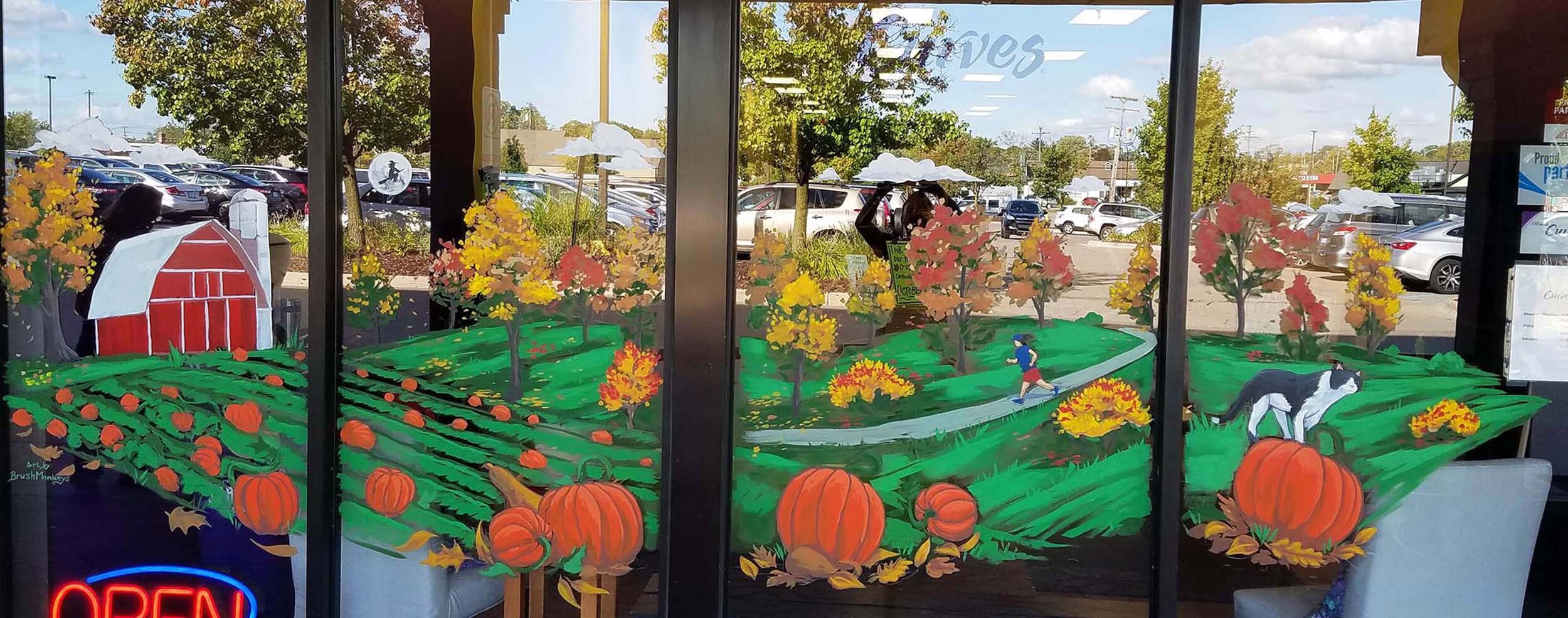 Vibrant autumn-themed window painting featuring pumpkins, a red barn, trees, a runner, and a cat in a pastoral scene.