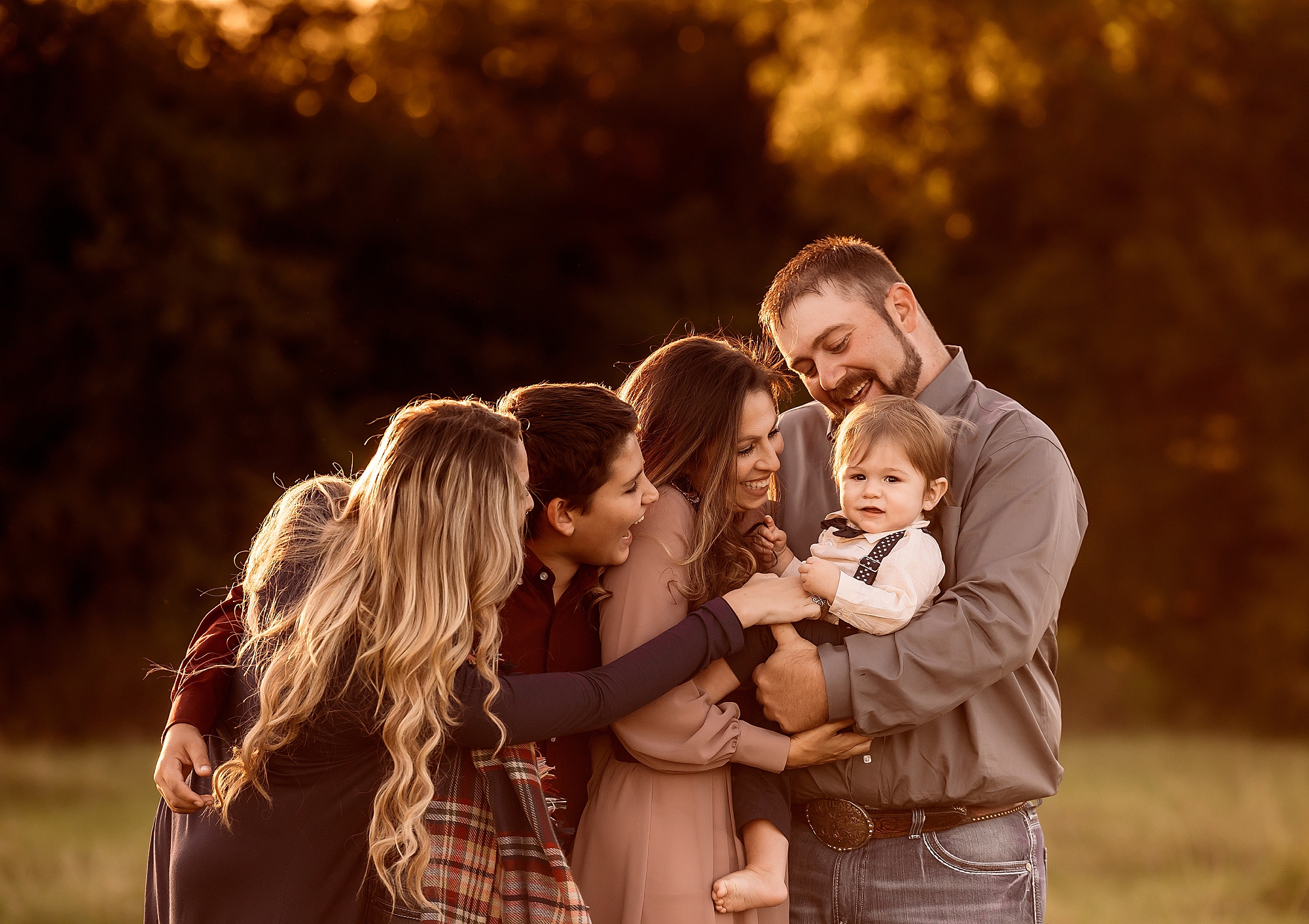 A joyful family of six hugging outdoors during golden hour, with warm sunlight filtering through trees in the background.