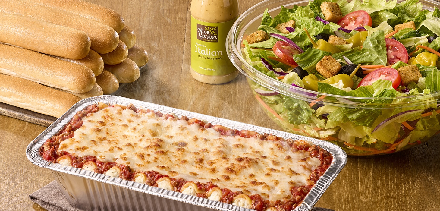 Olive Garden take-home meal featuring lasagna, breadsticks, Italian salad, and dressing on a wooden table.