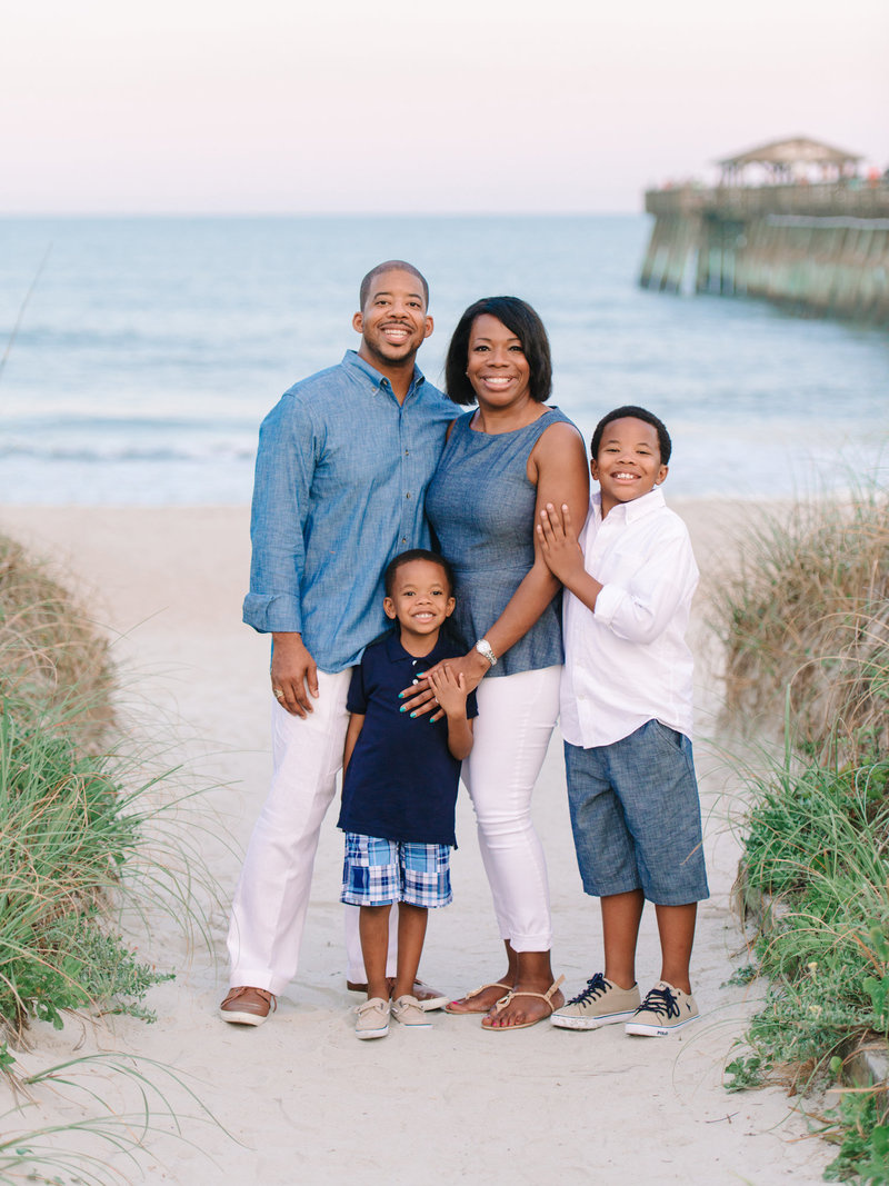A smiling Black family of four posing on a sandy beach path with ocean and pier in background at dusk.