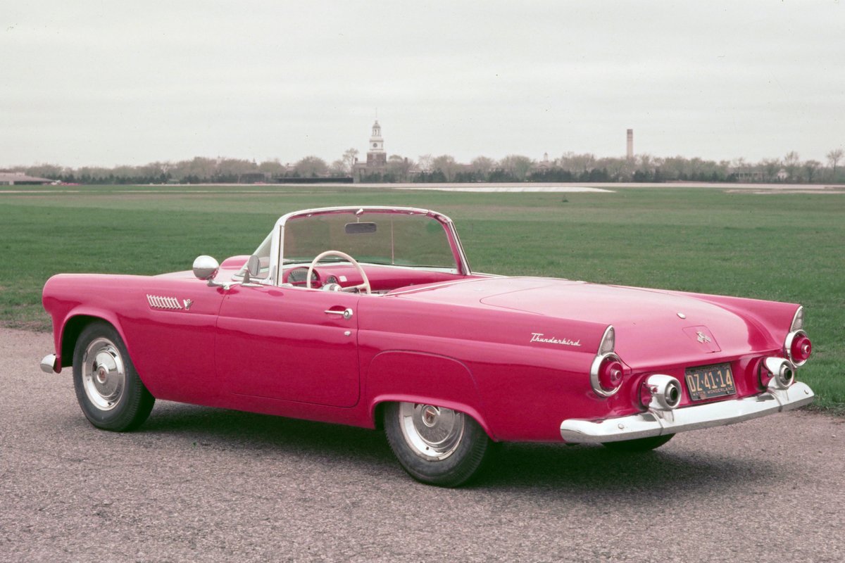 A vibrant red 1955 Ford Thunderbird convertible parked on gravel with a grassy field and historic building in the background.