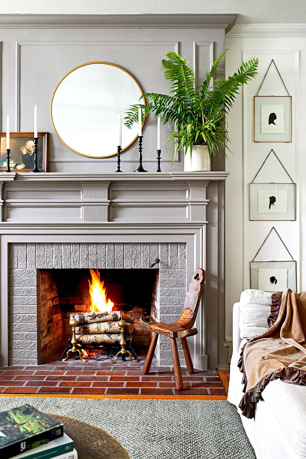Cozy living room with lit fireplace, gray mantel, round mirror, potted fern, vintage chair, and silhouette art on wall.