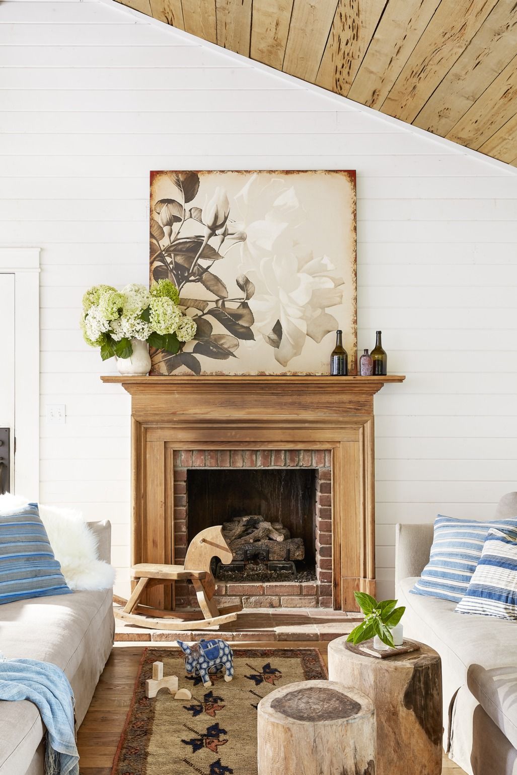 Rustic living room with wooden fireplace mantel, floral art, hydrangeas, striped pillows, and log side tables.