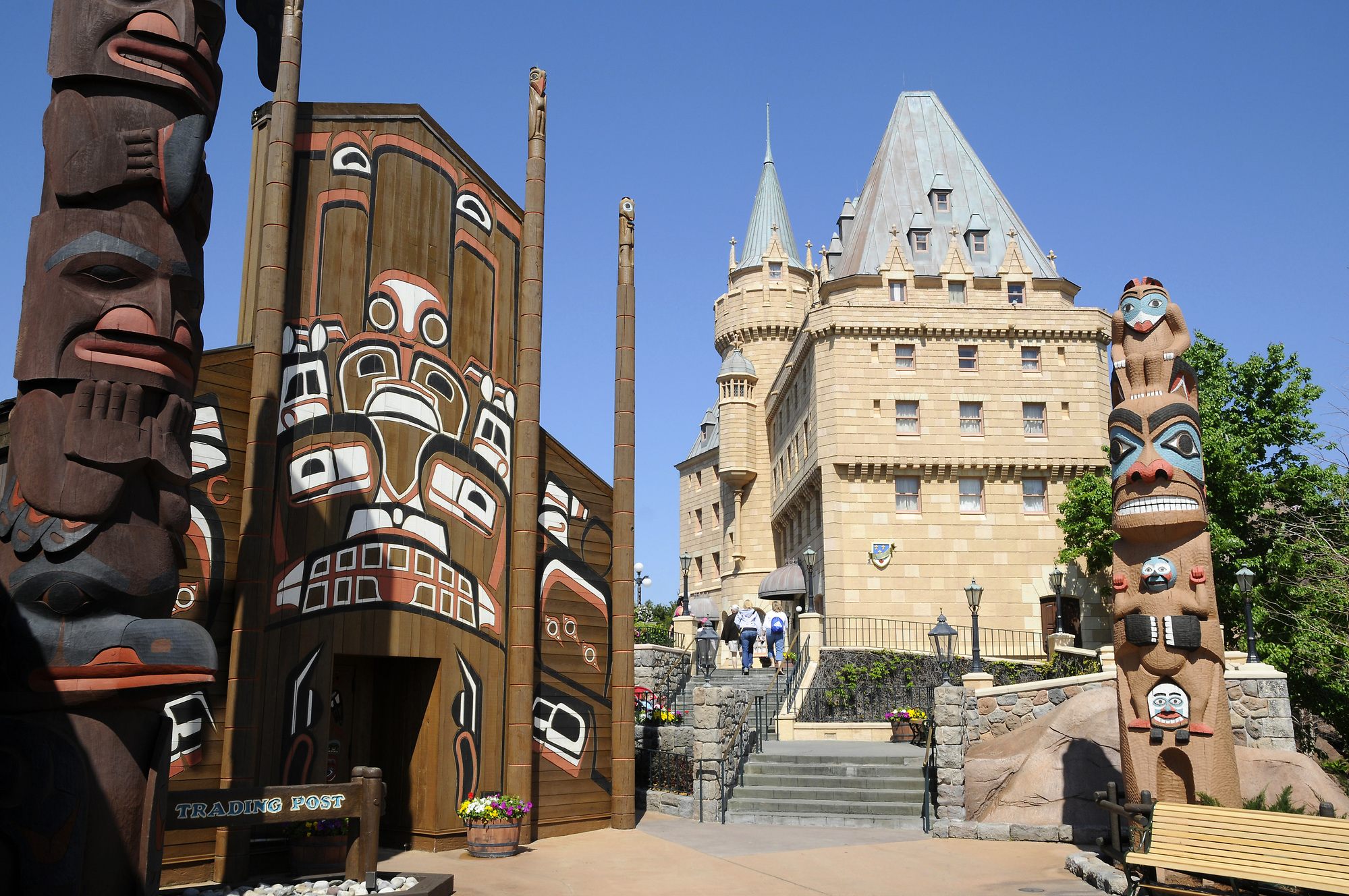 Totem poles and Pacific Northwest-style trading post beside a château-style castle under a clear blue sky.