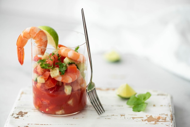 Fresh shrimp ceviche in a glass with lime, cilantro, tomato, cucumber, and a fork on a rustic white board.