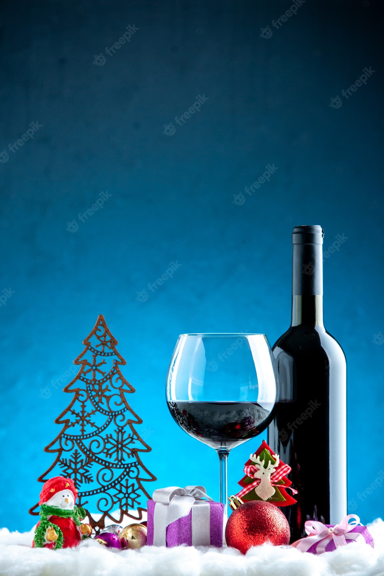 Festive holiday scene with red wine bottle and glass, decorative Christmas tree, snowman, gifts, and ornaments on snowy surface against blue backdrop.