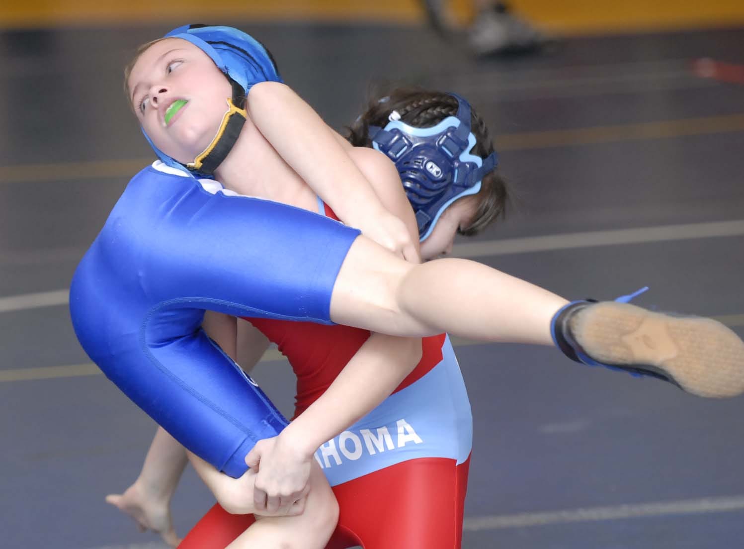 Two young female wrestlers in action-blue uniform executing a takedown on red-and-blue opponent during a match.
