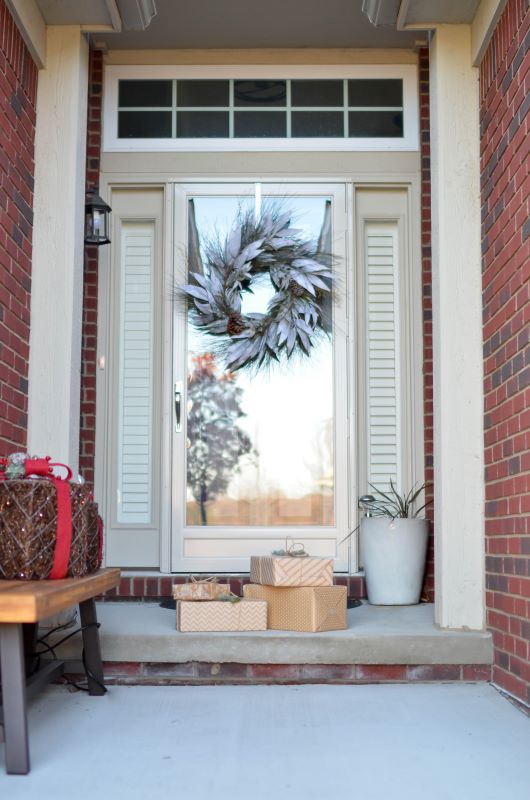Festive front porch with silver-gray wreath on glass door, wrapped gifts, and potted plant beside wooden bench.