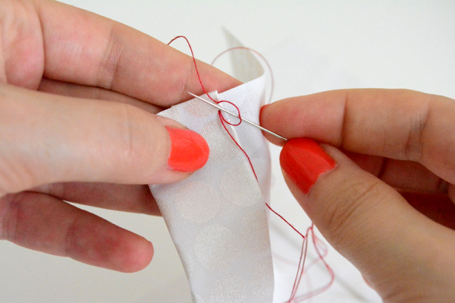 Close-up of hands with red nail polish hand-sewing white fabric using a needle and red thread.