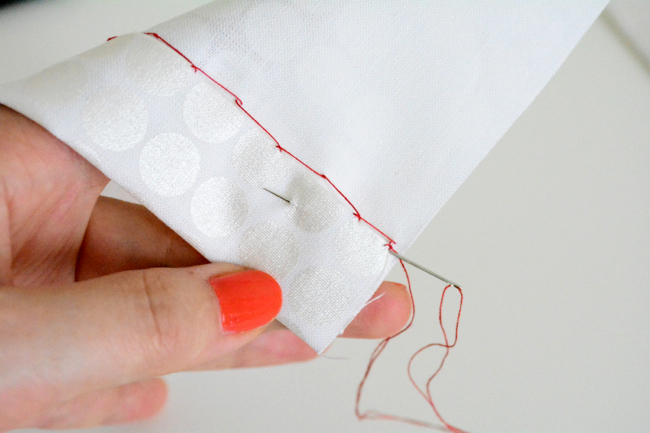 A hand with orange nail polish hand-stitching a white polka-dot fabric cuff with red thread using a needle.