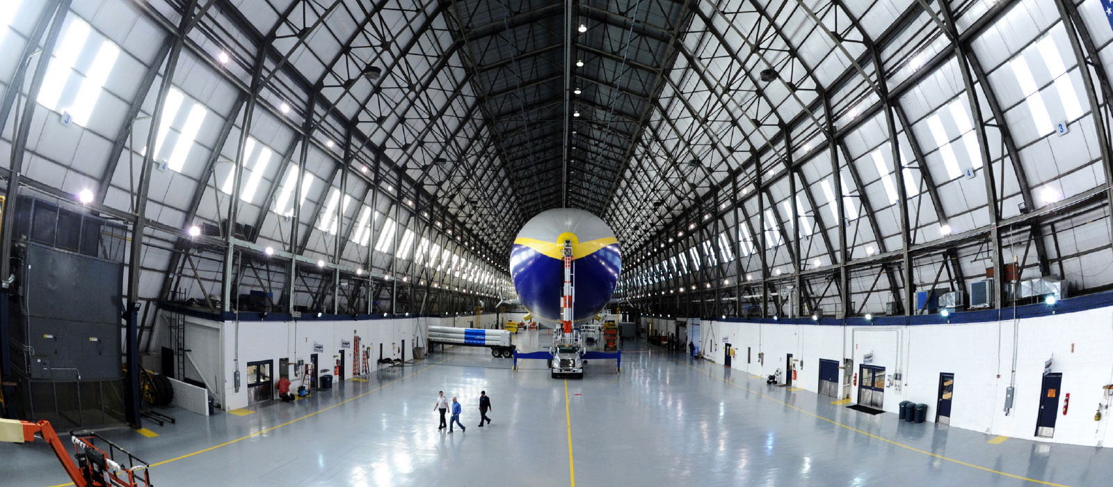 A massive blue-and-yellow aerostat suspended inside a high-ceiling hangar with workers and equipment below.