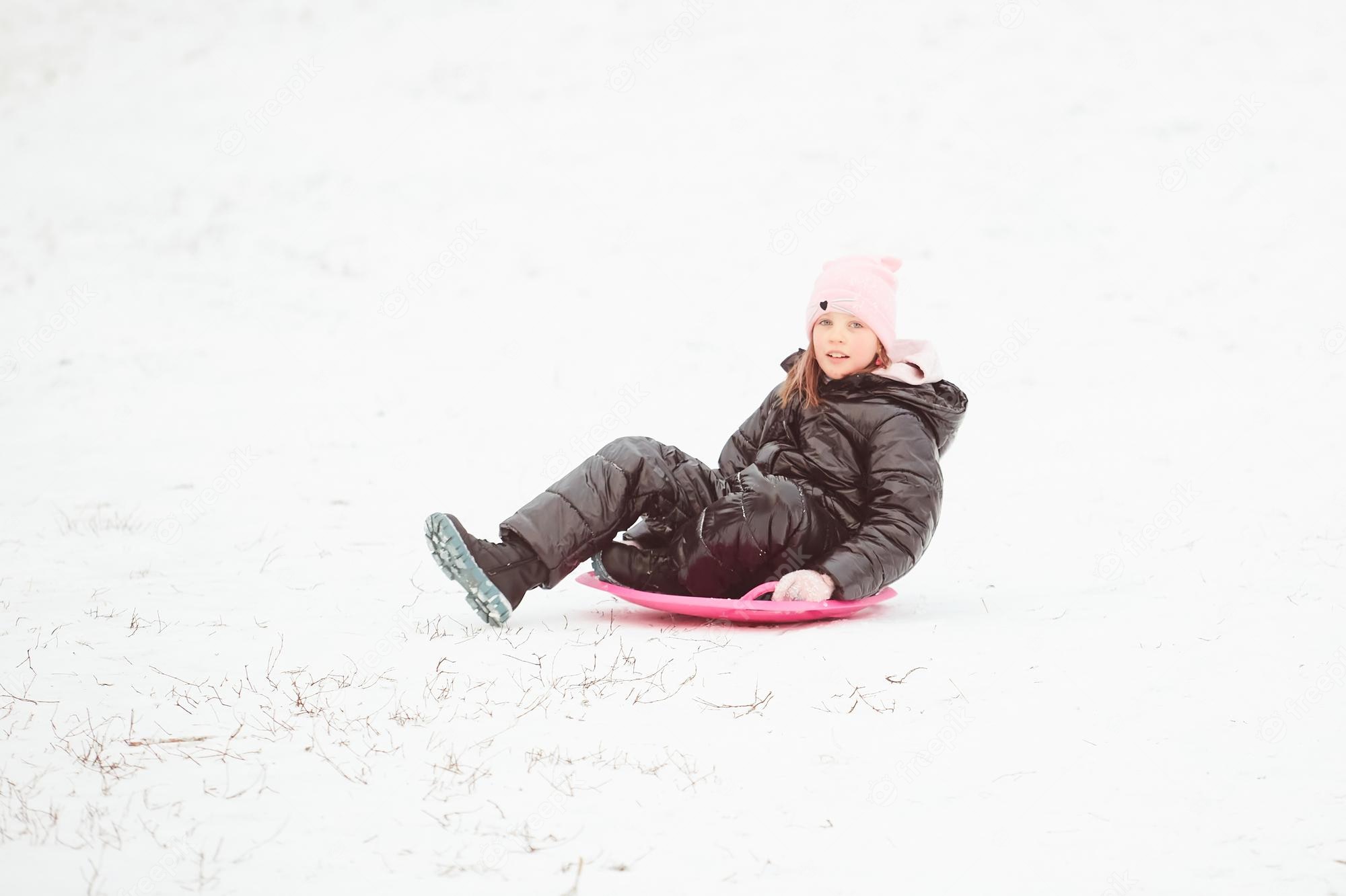 A young girl in a black winter coat and pink hat sits on a pink sled in the snow, smiling at the camera.