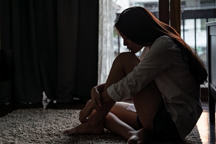 A young woman sits alone on a carpeted floor, head bowed in quiet contemplation near a window with soft light.