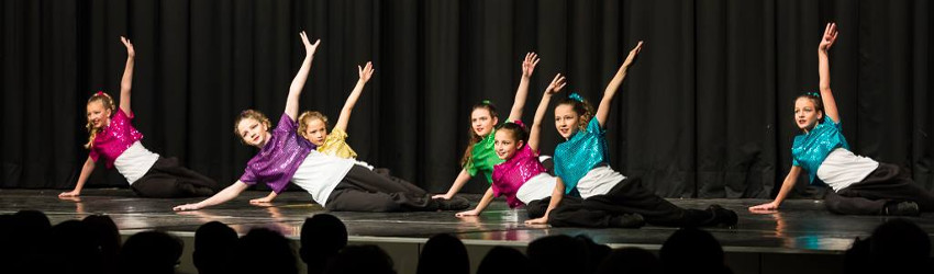 Seven young dancers in colorful sequined tops perform a synchronized floor pose on stage with black curtain backdrop.