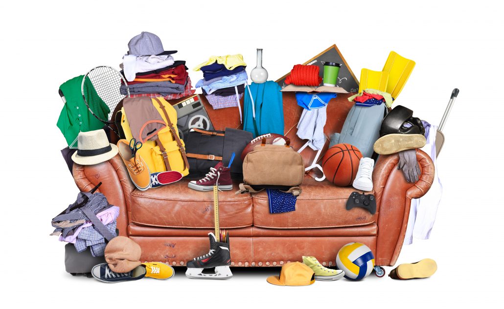 A cluttered brown leather sofa overflowing with clothes, sports gear, bags, shoes, and random household items against a white background.