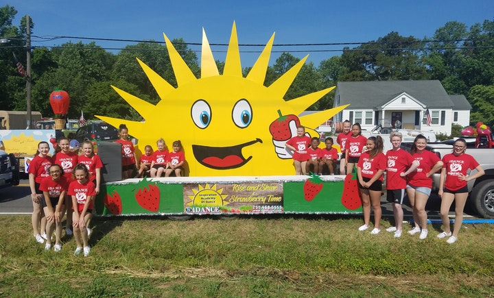 Group of smiling people in red shirts posing with a large yellow sun float decorated with strawberries and "Strawberry Time" signage.