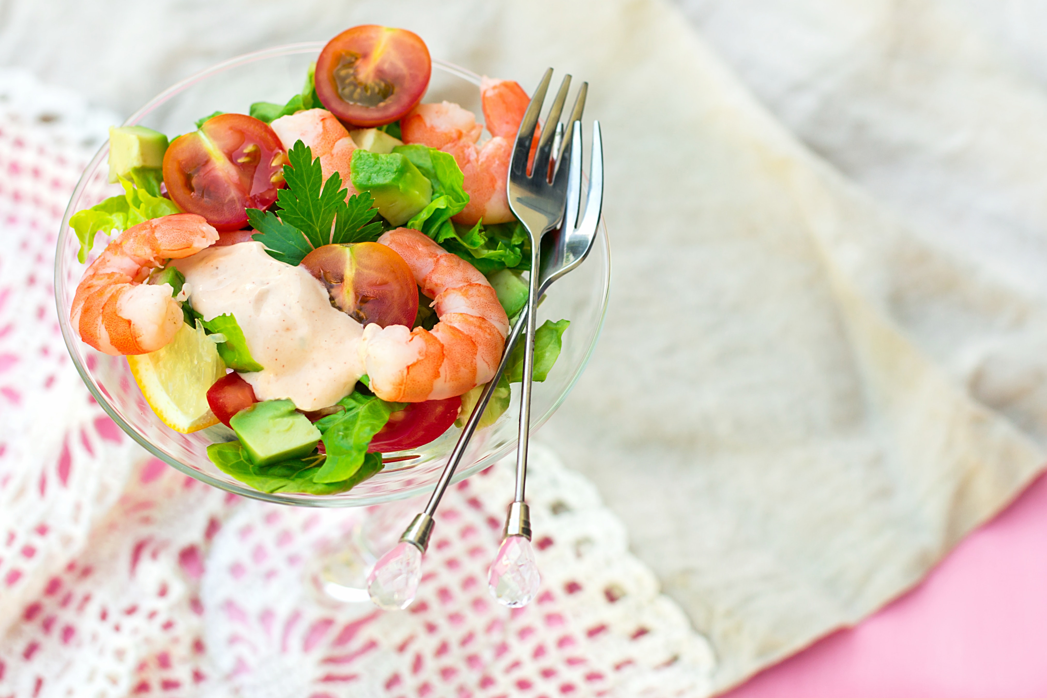 Fresh shrimp salad with cherry tomatoes, avocado, lettuce, lemon, and creamy dressing in a glass bowl with crystal-handled forks.
