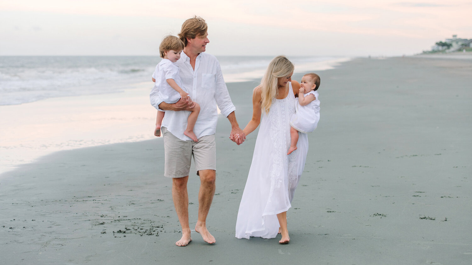 Family of four walking barefoot on a serene beach at dusk, dressed in white and light tones.