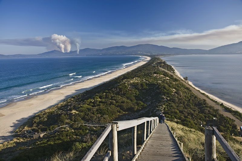 Wooden boardwalk descending a vegetated sand spit toward a beach, with distant smoke plume over mountains and ocean on both sides.