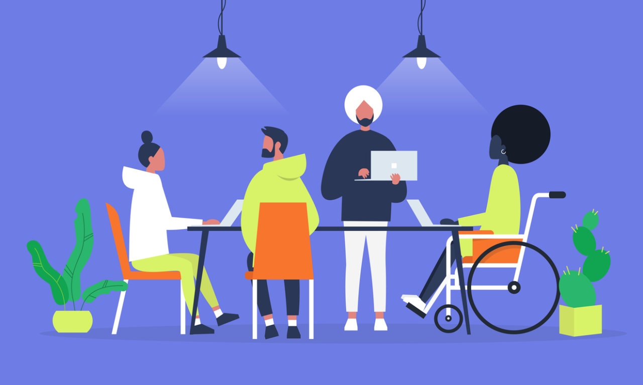 Diverse team collaborating at a table with laptops; one person uses a wheelchair, under modern pendant lights and potted plants.