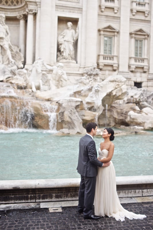 A newlywed couple embraces tenderly in front of Rome’s Trevi Fountain, with cascading water and classical sculptures in the background.