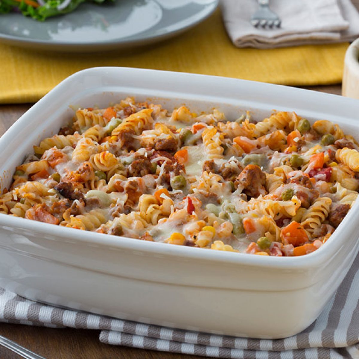 Baked pasta casserole with ground meat, peas, carrots, corn, and melted cheese in a white ceramic dish.